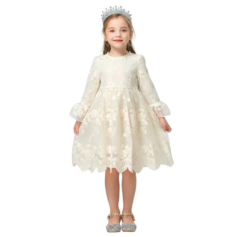 A young girl wearing a lace white dress with long sleeves and a scalloped hemline. She has sparkly silver shoes and a silver crown, standing against a plain white background.