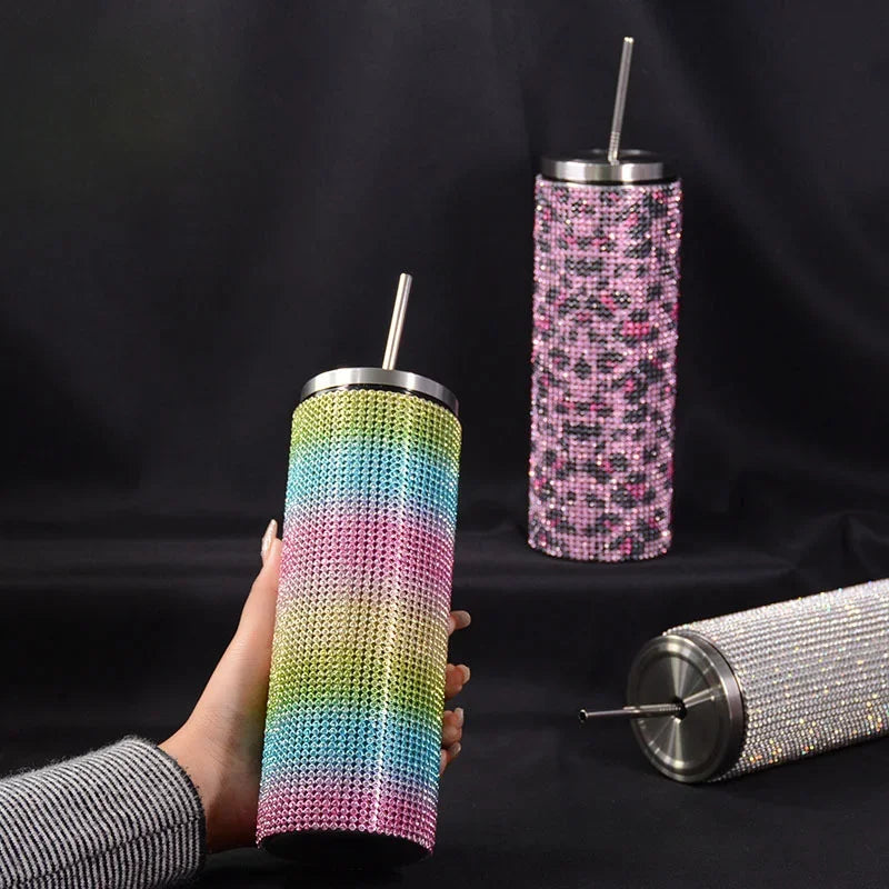 A hand holds a colorful rhinestone-covered tumbler with a metal straw. Two similar tumblers, one pink leopard print and one silver, are in the background against a black backdrop.