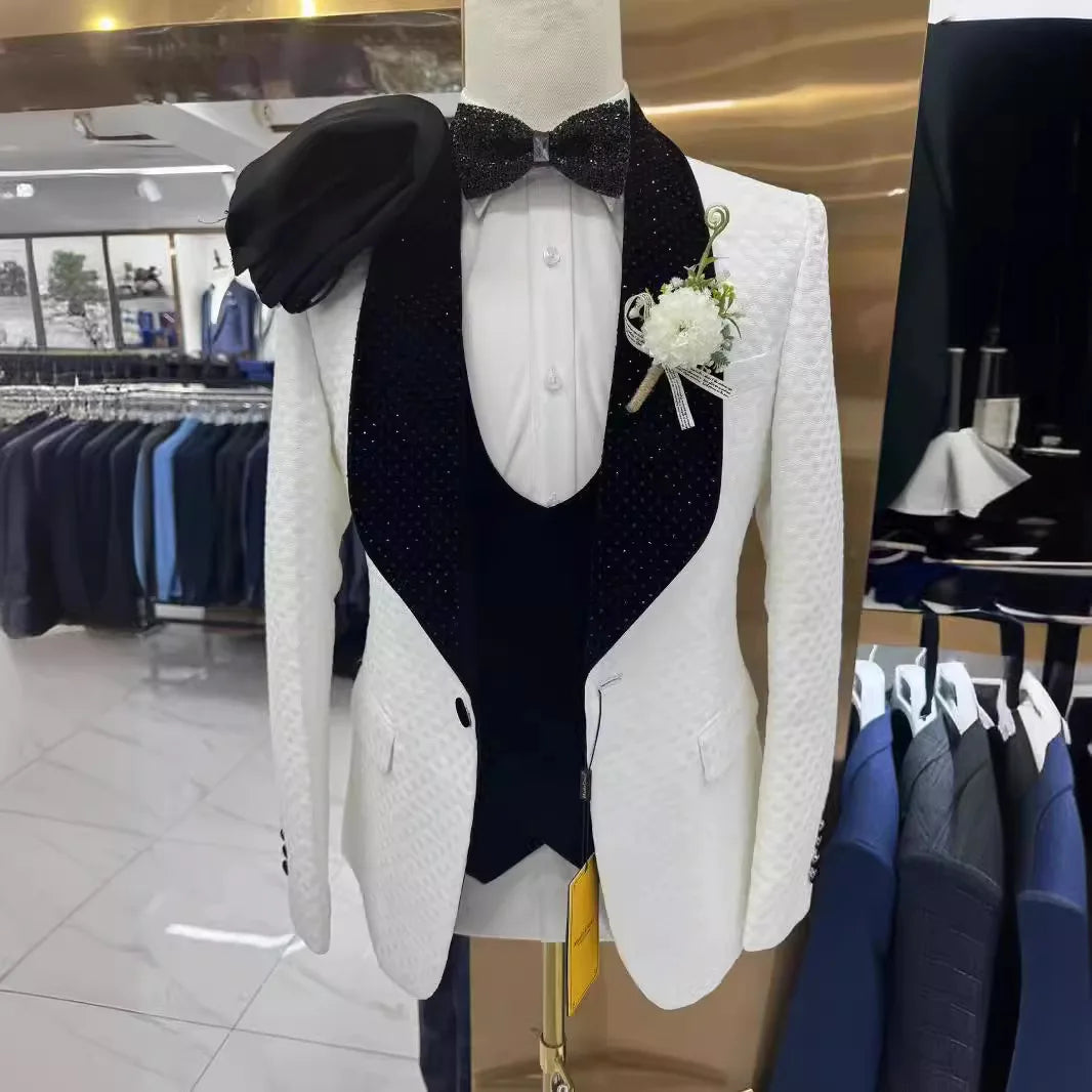 A mannequin displays a white textured tuxedo with a black, sparkly lapel and bow tie, white shirt, and boutonniere. A black hat rests on the shoulder. Suits hang in the background.