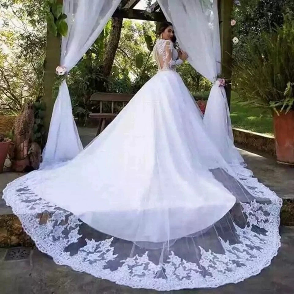 Bride in a white gown stands under a decorated pergola. The dress has a long, lacy train, and the setting is a lush garden with potted plants and white draped curtains.