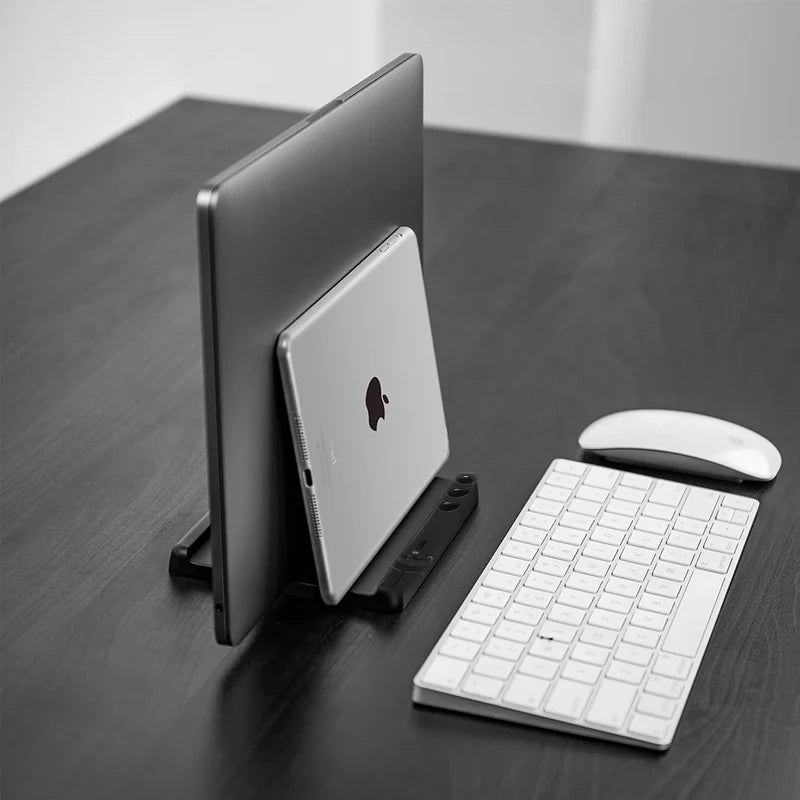 A laptop and tablet are vertically stored in a black stand on a desk. A wireless keyboard and mouse are placed beside them, all in a minimalist setup.