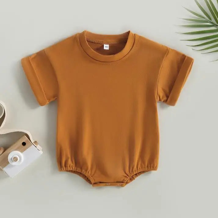 Rust-colored baby romper with short sleeves, laid flat on a light surface. A small wooden toy camera sits on the left, and a green palm leaf is on the right.