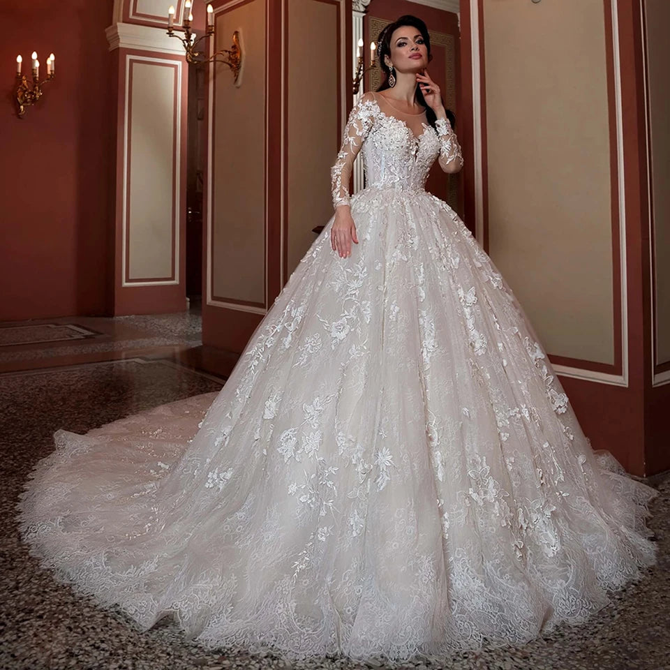 Woman in an ornate, long-sleeved lace wedding gown stands in an elegant hallway with chandelier lighting. The dress features floral patterns and a voluminous skirt with a long train.