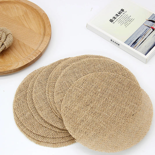A stack of round burlap coasters on a table, next to a wooden plate and an open book with a beach scene cover. The items are arranged on a white surface.