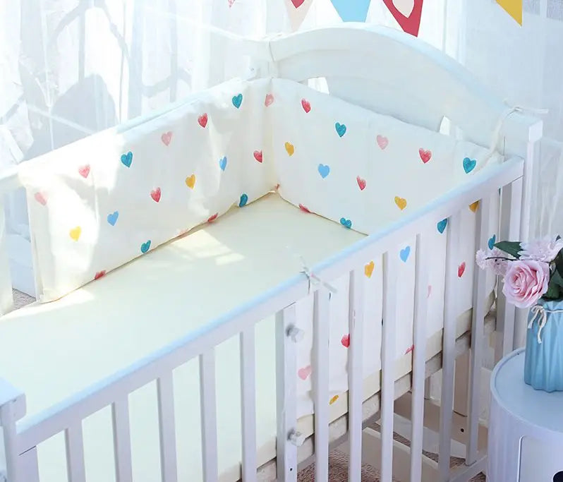 White baby crib with heart-patterned bumpers in pastel blue, pink, yellow, and red. Sunlight filters through sheer curtains. Pink flowers decorate a nearby blue table.