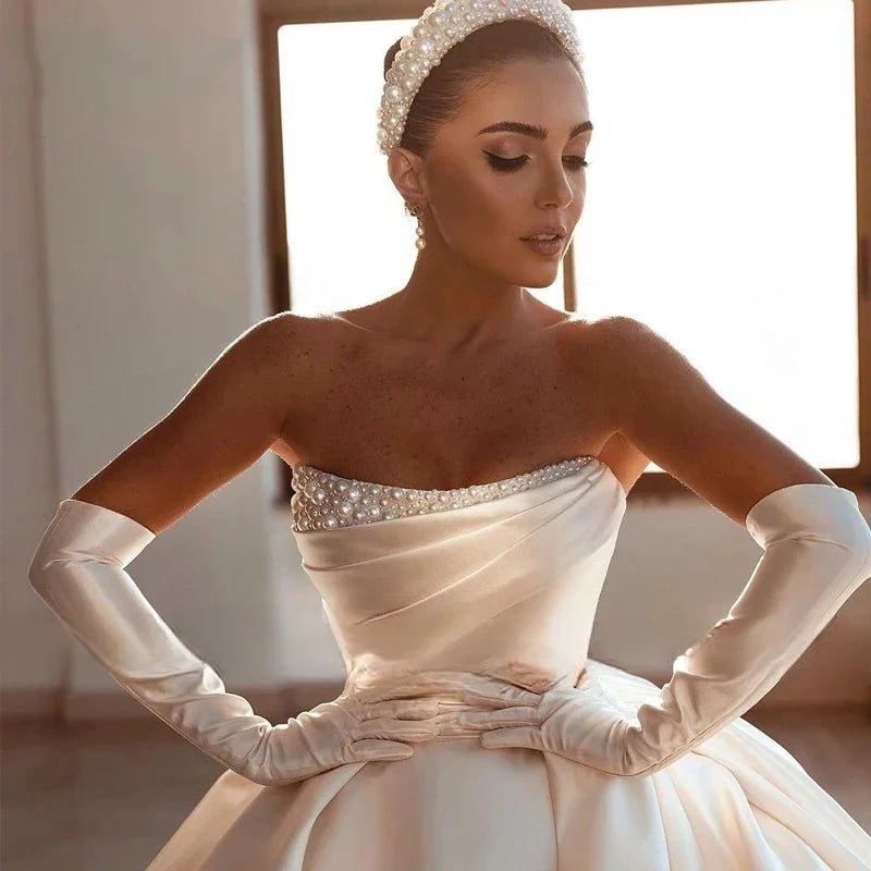Bride in an elegant, strapless satin gown with pearl embellishments, wearing long gloves and a pearl headband, stands with hands on hips in a softly lit room.