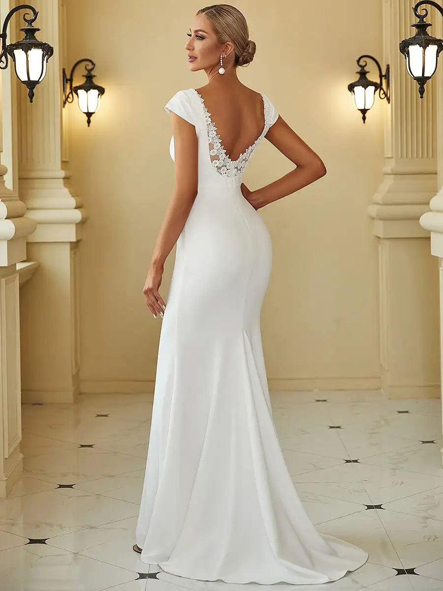 Woman in an elegant white gown with a deep V-back and lace detailing stands in a marble hallway. Her hair is styled in a bun, complemented by pearl earrings, under classic wall lamps.