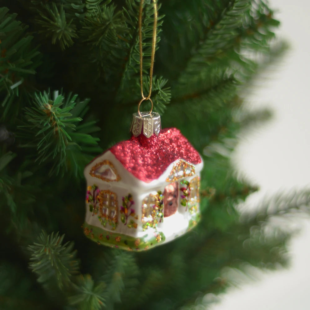 A Christmas tree ornament depicting a glittery house with a red roof and colorful details hangs from a lush green tree branch.