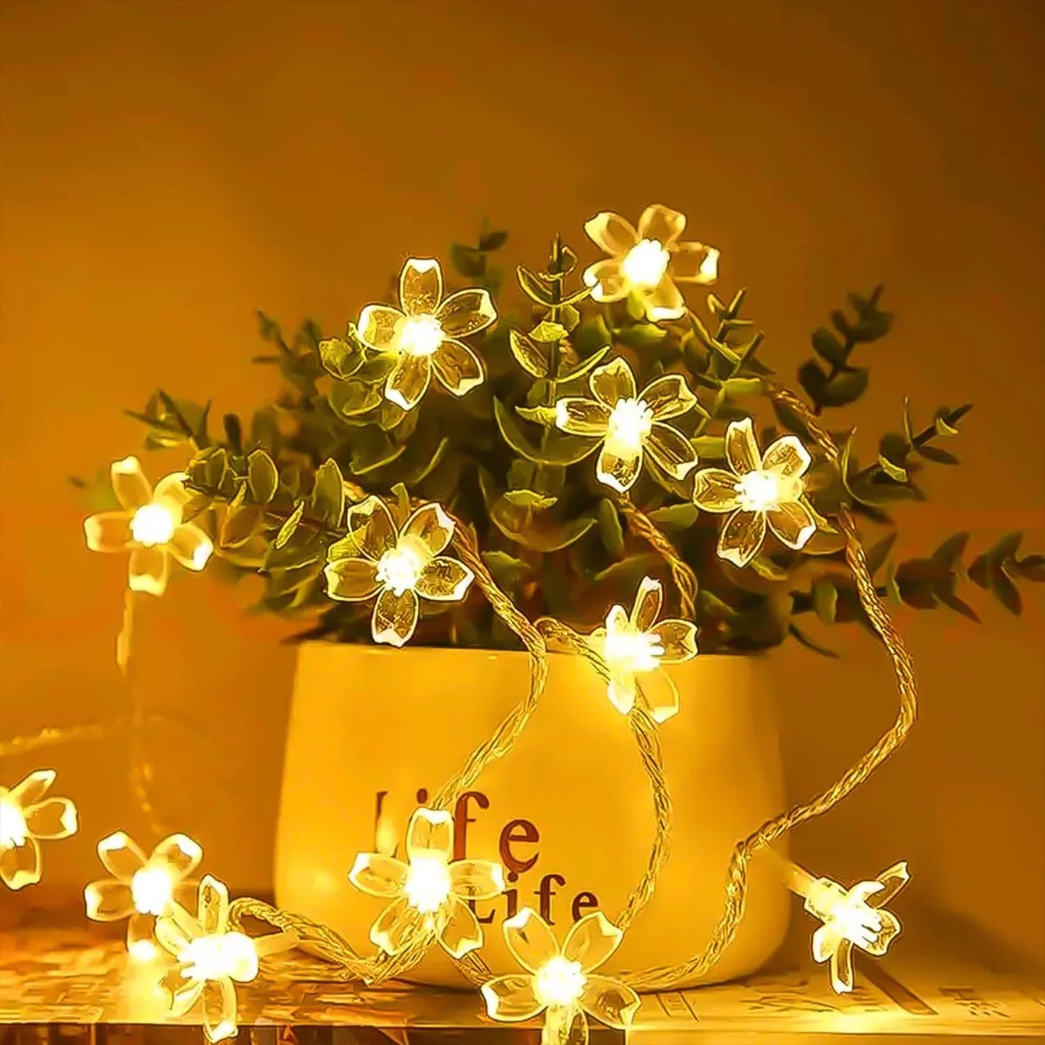 String lights shaped like flowers illuminate a small plant in a pot with "Life" text. The warm yellow glow creates a cozy ambiance against a soft, blurred background.