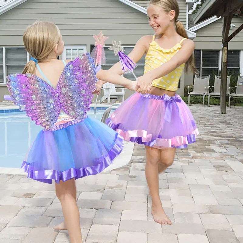 Two girls in colorful tutus and fairy wings play joyfully by a backyard pool, holding star-shaped wands. One wears blue wings, the other a yellow polka dot top.