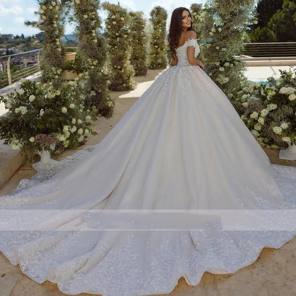 A bride in an elegant, off-shoulder wedding gown with a long train stands on a floral-decorated terrace, gazing back with a smile amidst lush greenery and clear skies.