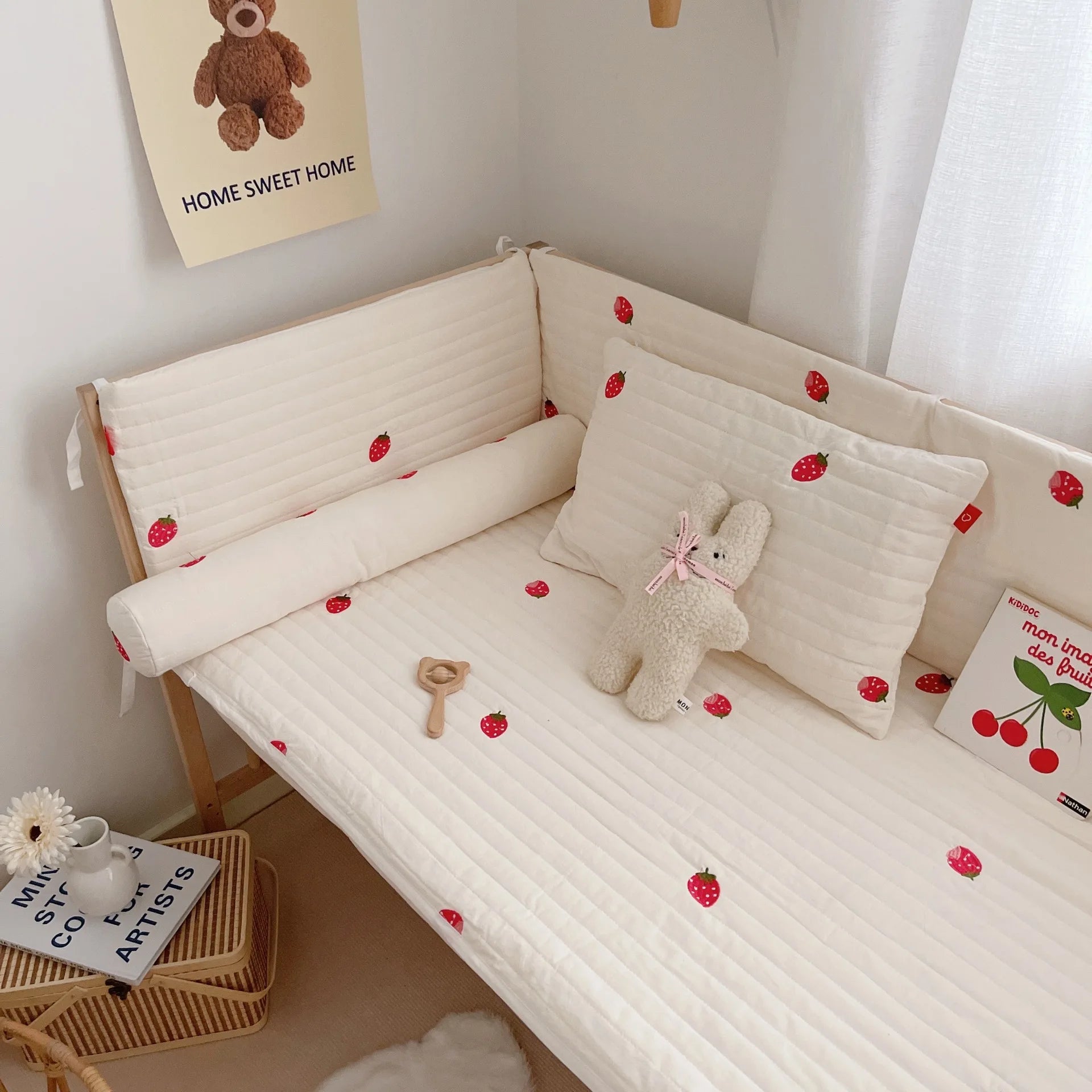 Cozy child's bed with strawberry-patterned bedding, a plush toy, and a wooden rattle. Nearby, a small table holds a vase, flower, and books. A teddy bear poster decorates the wall.