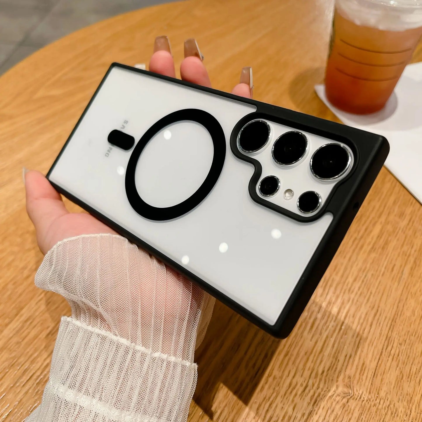 A hand holding a smartphone with a unique white and black case featuring a large circle and multiple camera lenses. A drink is on the wooden table in the background.