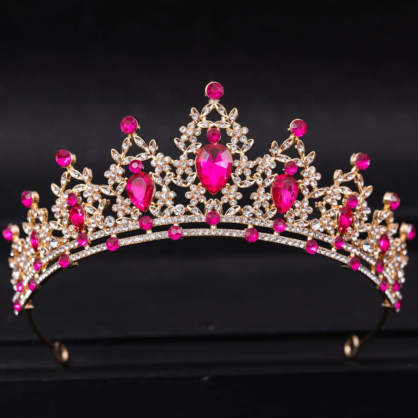 A gold tiara adorned with sparkling white crystals and vivid pink gemstones in an ornate design, set against a black background.