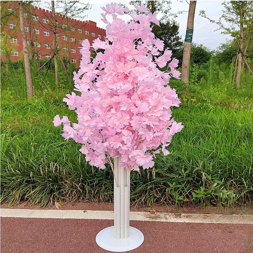 Pink artificial tree with dense blossoms in a tall, white vase set on a paved path. Green grass and trees, with a red brick building in the background.