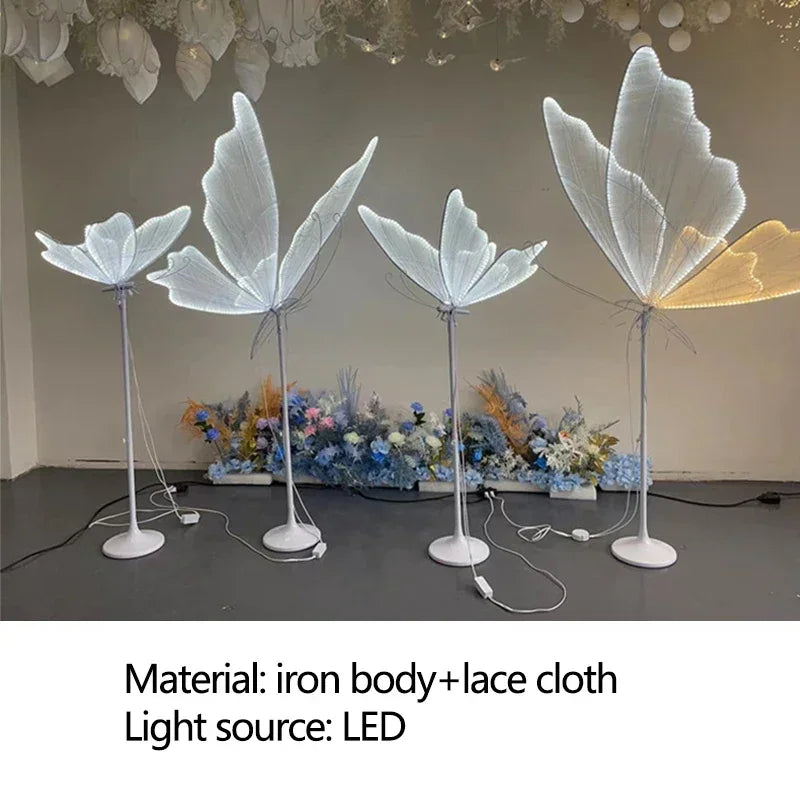 Four LED floor lamps with large, white, leaf-like shades made from lace cloth and supported by iron stands are displayed indoors, with decorative flowers in the background.