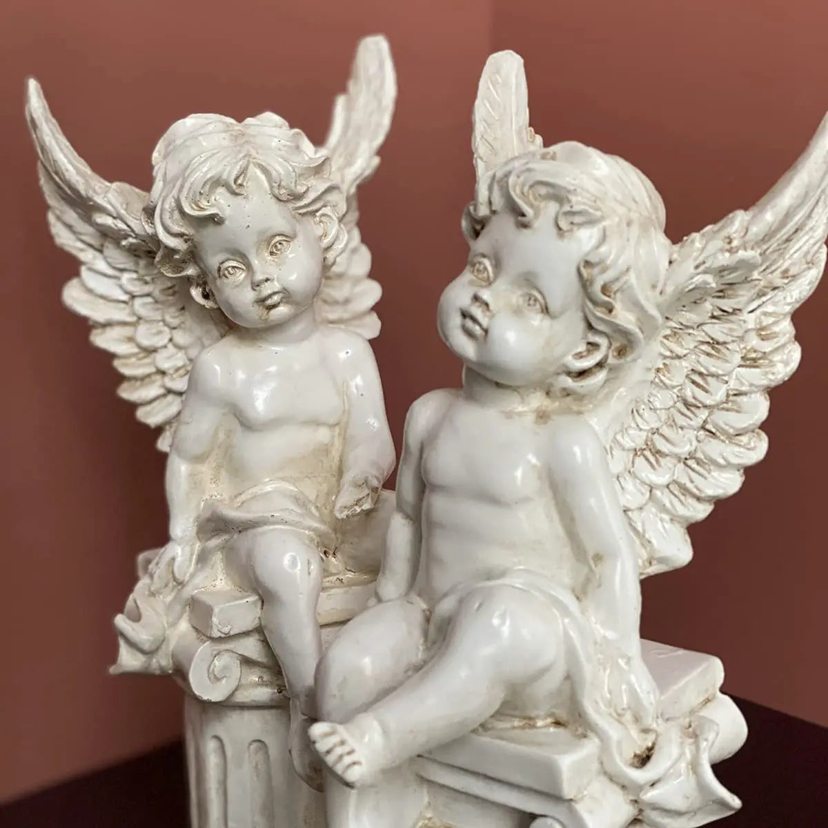 Two white ceramic cherub statues with detailed wings sit on ornate columns against a warm brown background.