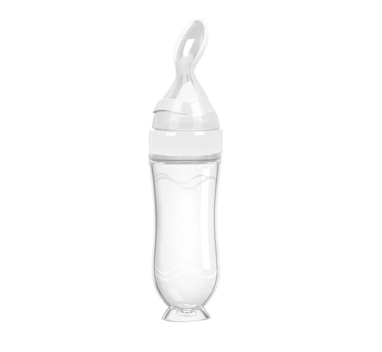 Transparent baby bottle with a white silicone spoon attachment at the top, designed for easy feeding. The bottle has a sleek, ergonomic shape on a white background.