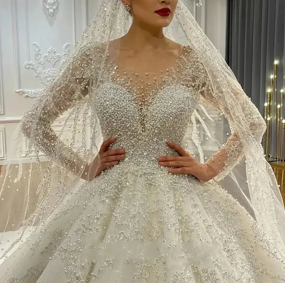 Bride in an ornate wedding gown adorned with pearls and intricate lace, standing in an elegant room with her hands on her waist, wearing a detailed veil.