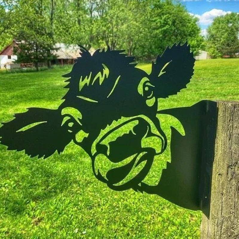 Metal cow cutout mounted on a wooden post in a green field. Trees and a barn are in the background under a blue sky with clouds.