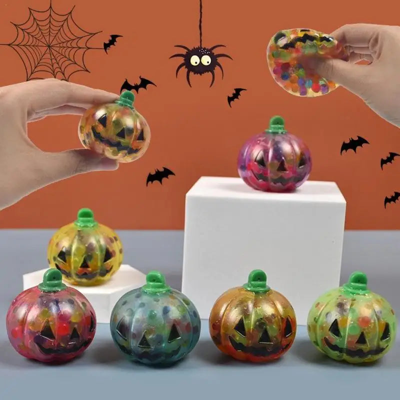 Colorful, translucent pumpkin toys with jack-o'-lantern faces filled with beads, held by hands. Halloween-themed background with hanging spider, web, and bats.