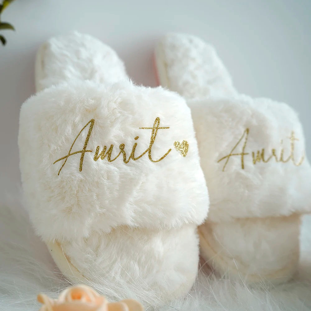 White fluffy slippers with gold embroidery spelling "Amrit" and a heart. The slippers are placed on a soft surface, with a faint hint of a peach-colored rose in the foreground.