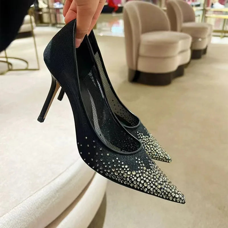 A close-up of a hand holding a pair of black high-heeled shoes adorned with sparkling rhinestones. The background features beige armchairs and a soft carpeted floor.