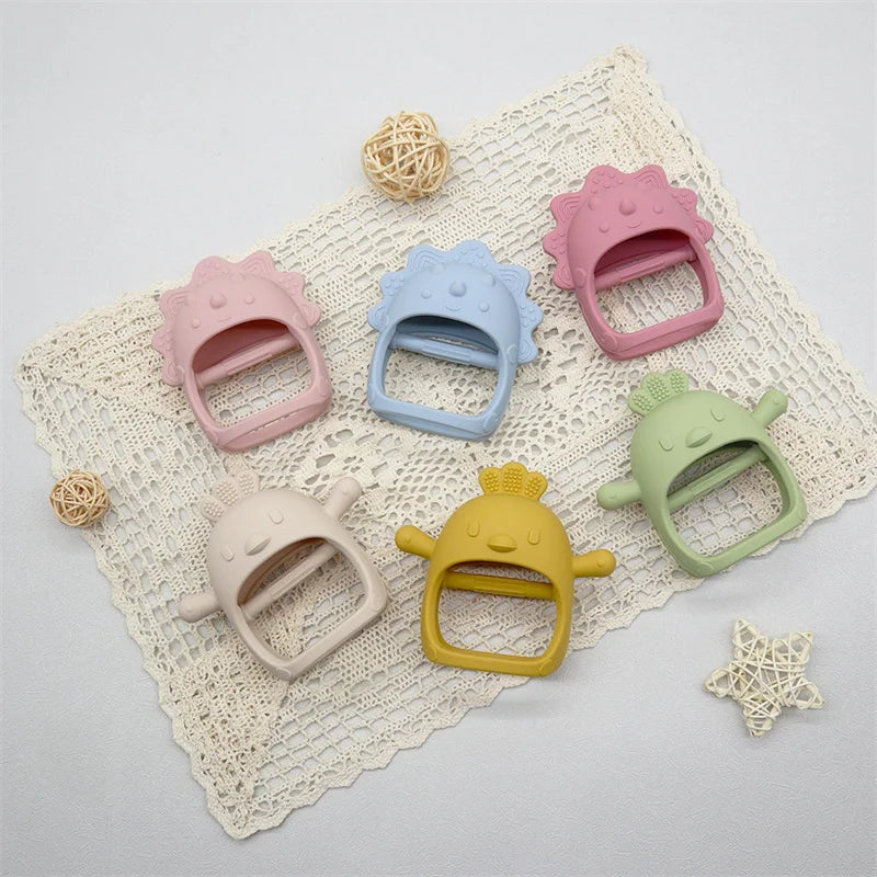 Six colorful, cartoon-style silicone teethers shaped like cheerful animals are arranged on a lace doily, surrounded by small decorative wicker balls and a star.