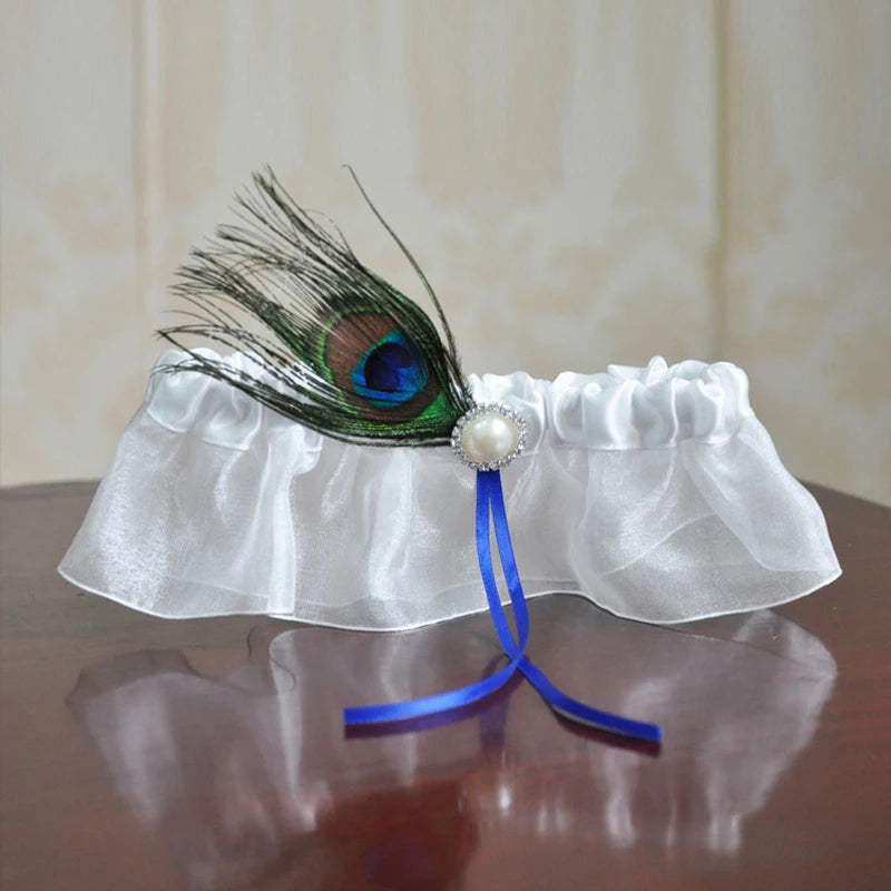 White satin garter with a peacock feather detail, adorned with a pearl and blue ribbon, placed on a reflective wooden surface.