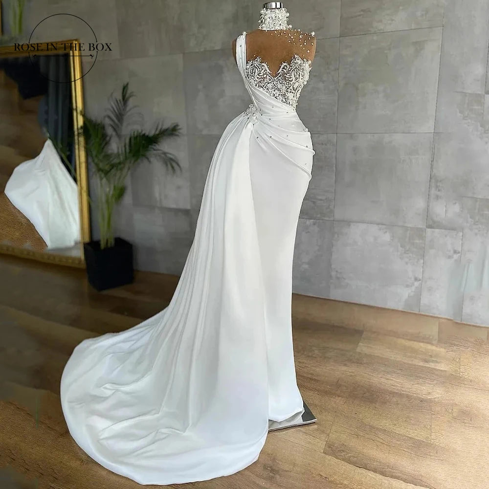 Elegant white dress on a mannequin with a sheer embellished lace bodice, draped one-shoulder design, and long flowing train. Set against a simple interior with wooden floors.