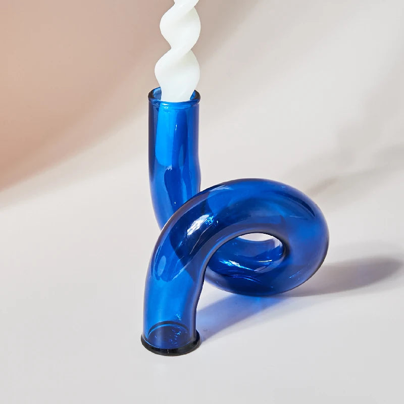A vibrant blue curved glass candle holder with a unique loop design holds a twisted white candle, set against a neutral background.
