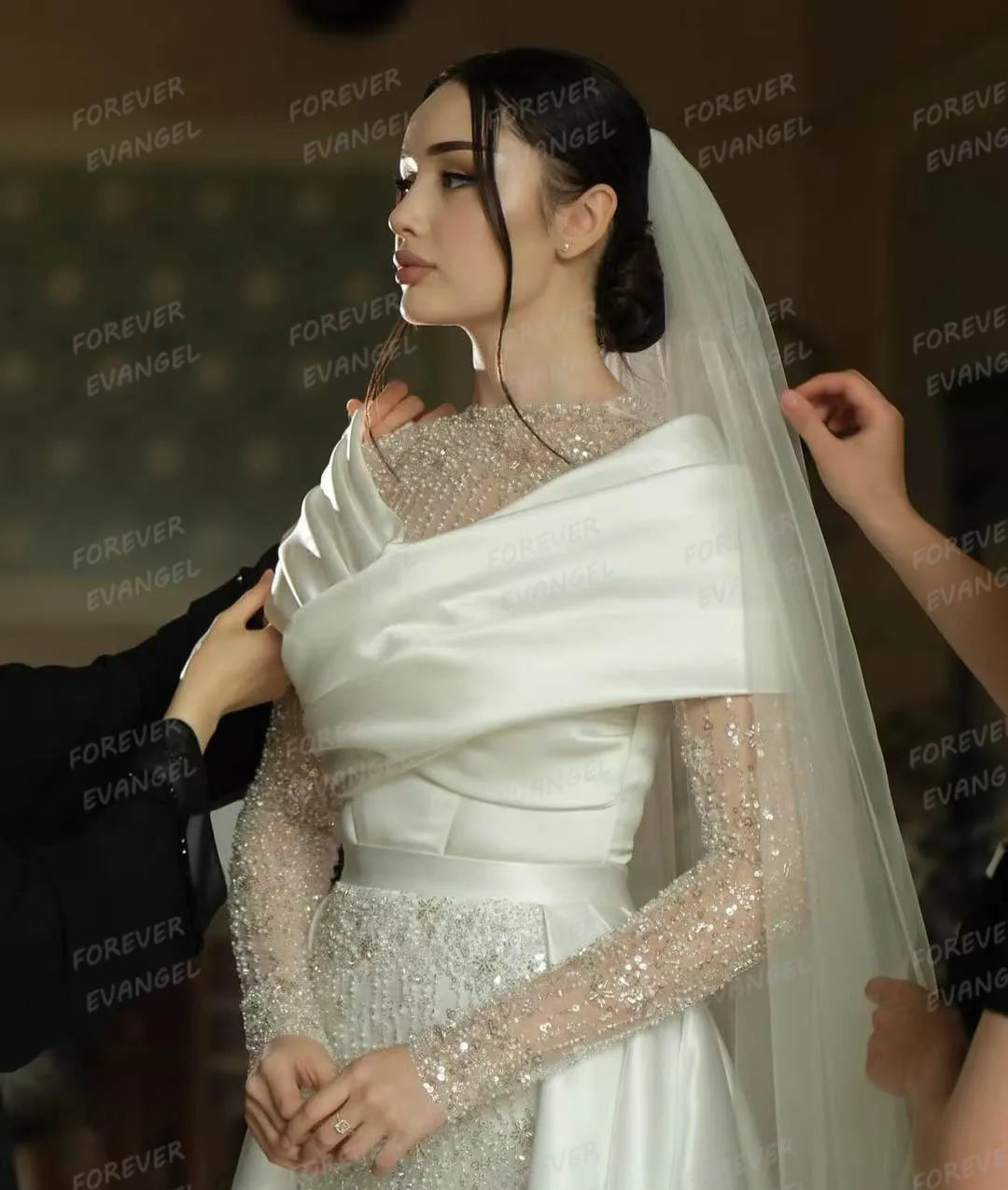 A bride wearing a sparkling beaded wedding dress with an elegant, draped white satin wrap. Her hair is styled in a bun adorned with a long veil, as hands adjust her attire.