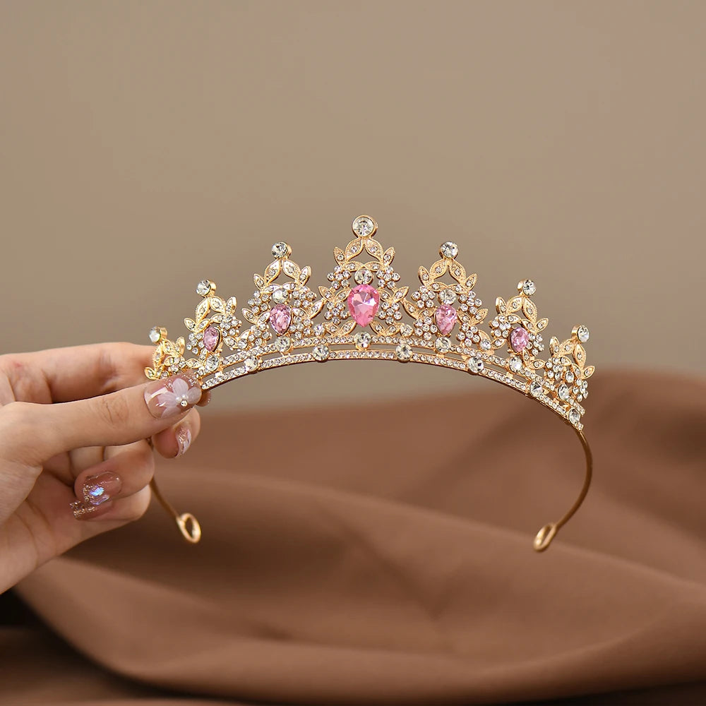 A hand holds a gold tiara adorned with sparkling clear and pink gemstones, set against a soft brown background. The tiara has intricate floral and heart designs.