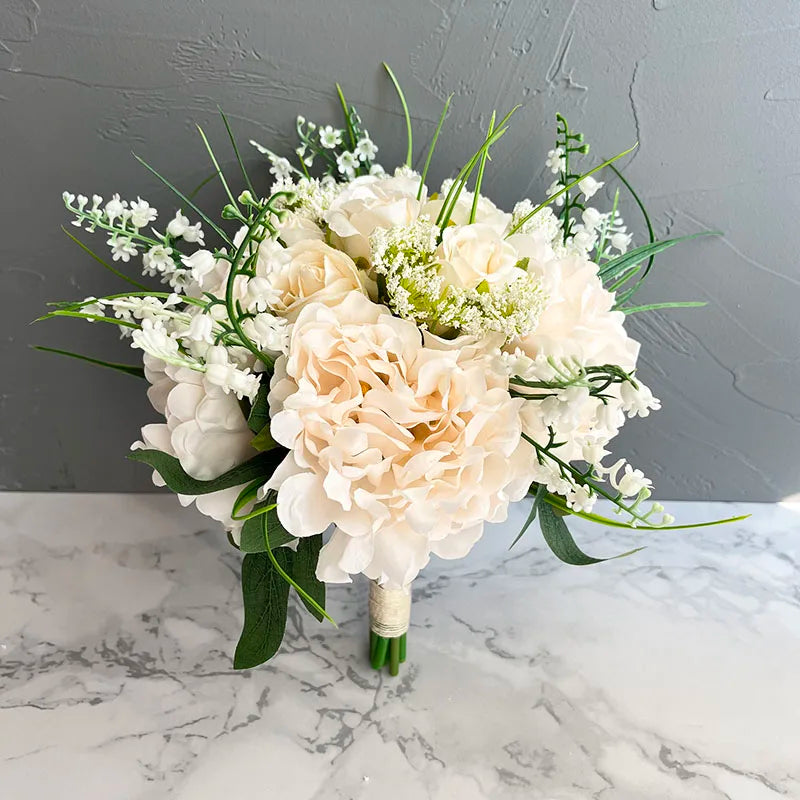 A bouquet of cream peonies, white roses, and greenery against a gray textured wall. The flowers are wrapped with a white ribbon and placed on a marble surface.