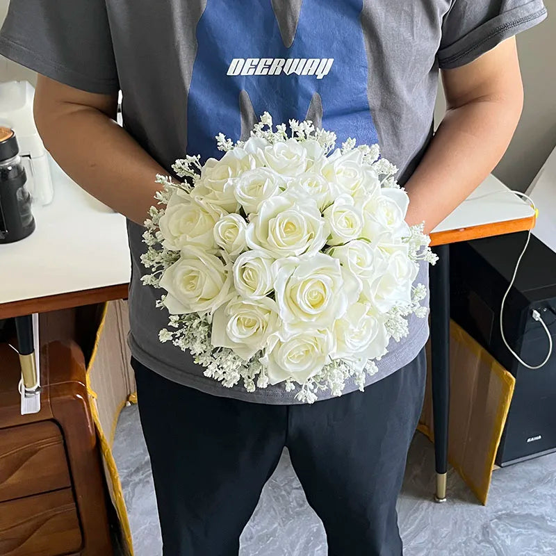 Person in a gray t-shirt holds a bouquet of white roses and small white flowers, standing indoors next to a table with a cup, on a gray floor.