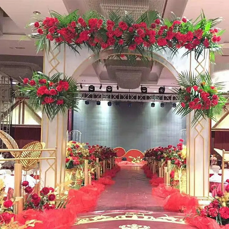 Elegant venue decorated with a gold and white arch adorned with red roses and green foliage, leading to an aisle flanked by red flower arrangements and soft lighting.