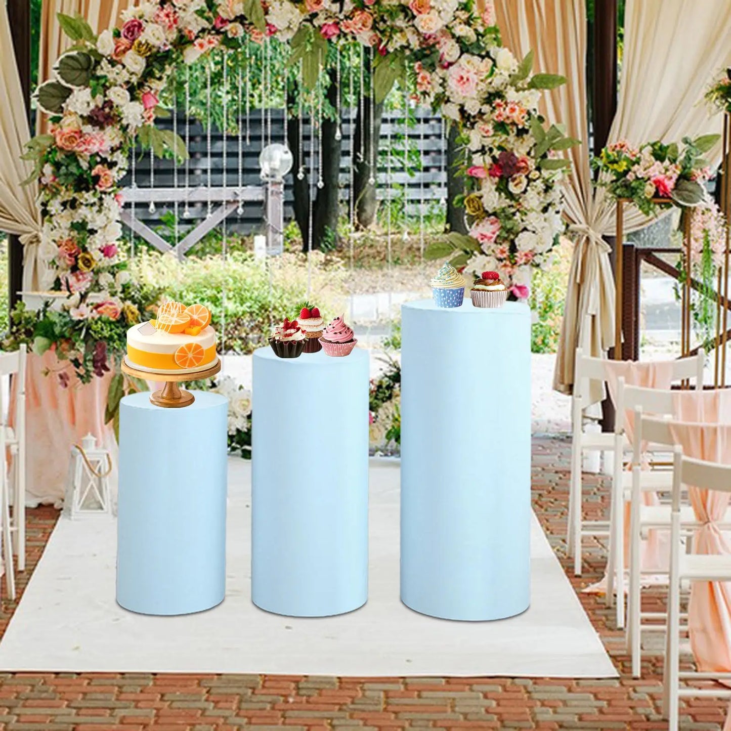 Wedding scene with a floral arch and three blue pedestals; a cake and cupcakes are displayed. White chairs line the aisle, and soft drapes add elegance to the decor.