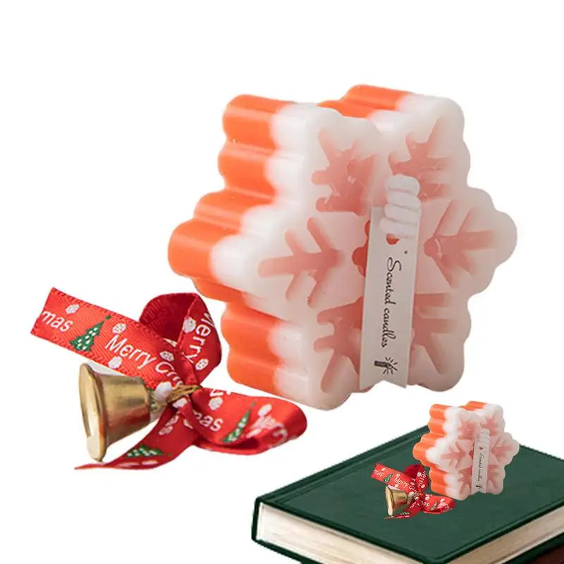 Snowflake-shaped candle with orange and white layers, featuring a Christmas-themed ribbon and bell. Displayed on a green book with a "Merry Christmas" tag.