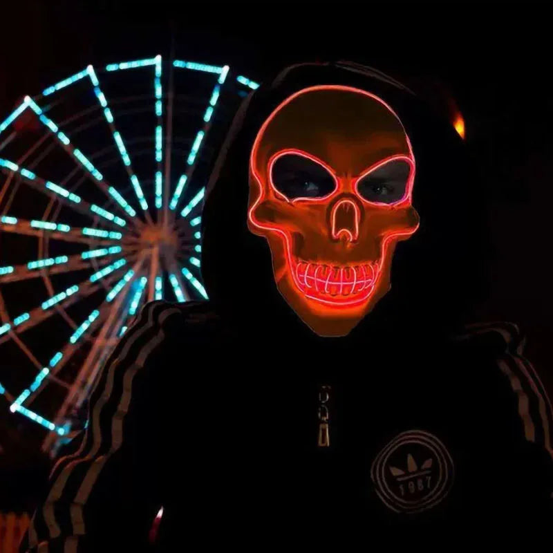 A person wearing an illuminated skull mask and a hooded jacket stands in front of a brightly lit Ferris wheel at night.