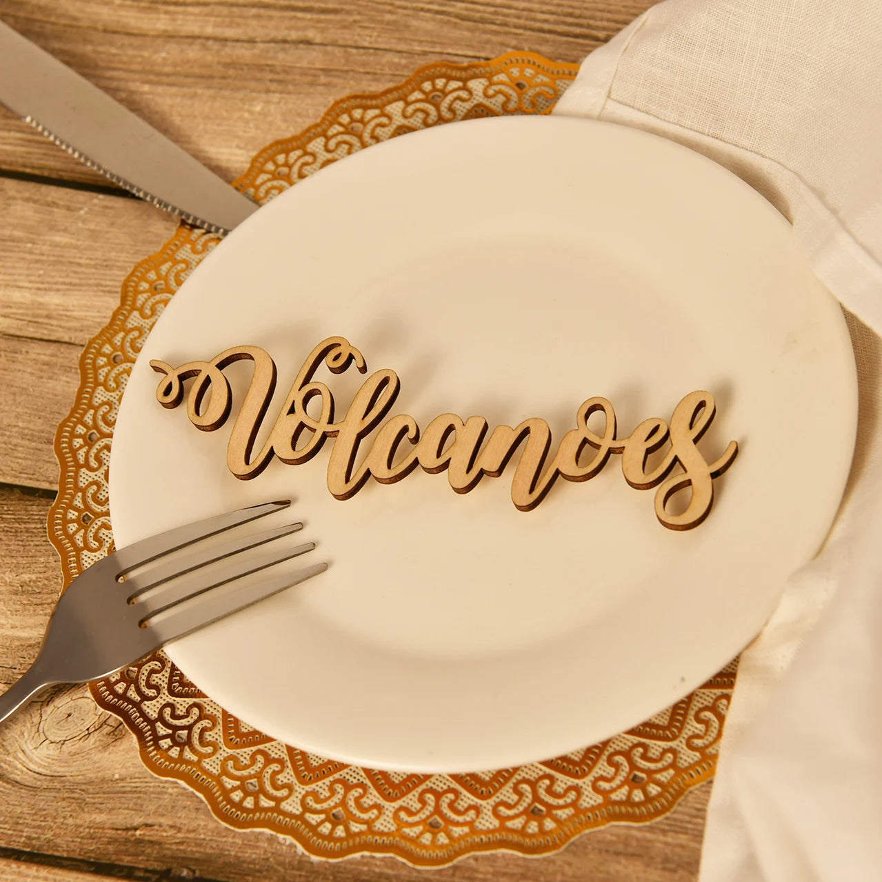 A wooden cutout of the word "Volcanoes" placed on a white plate, with a gold doily, fork, and knife on a wooden table.