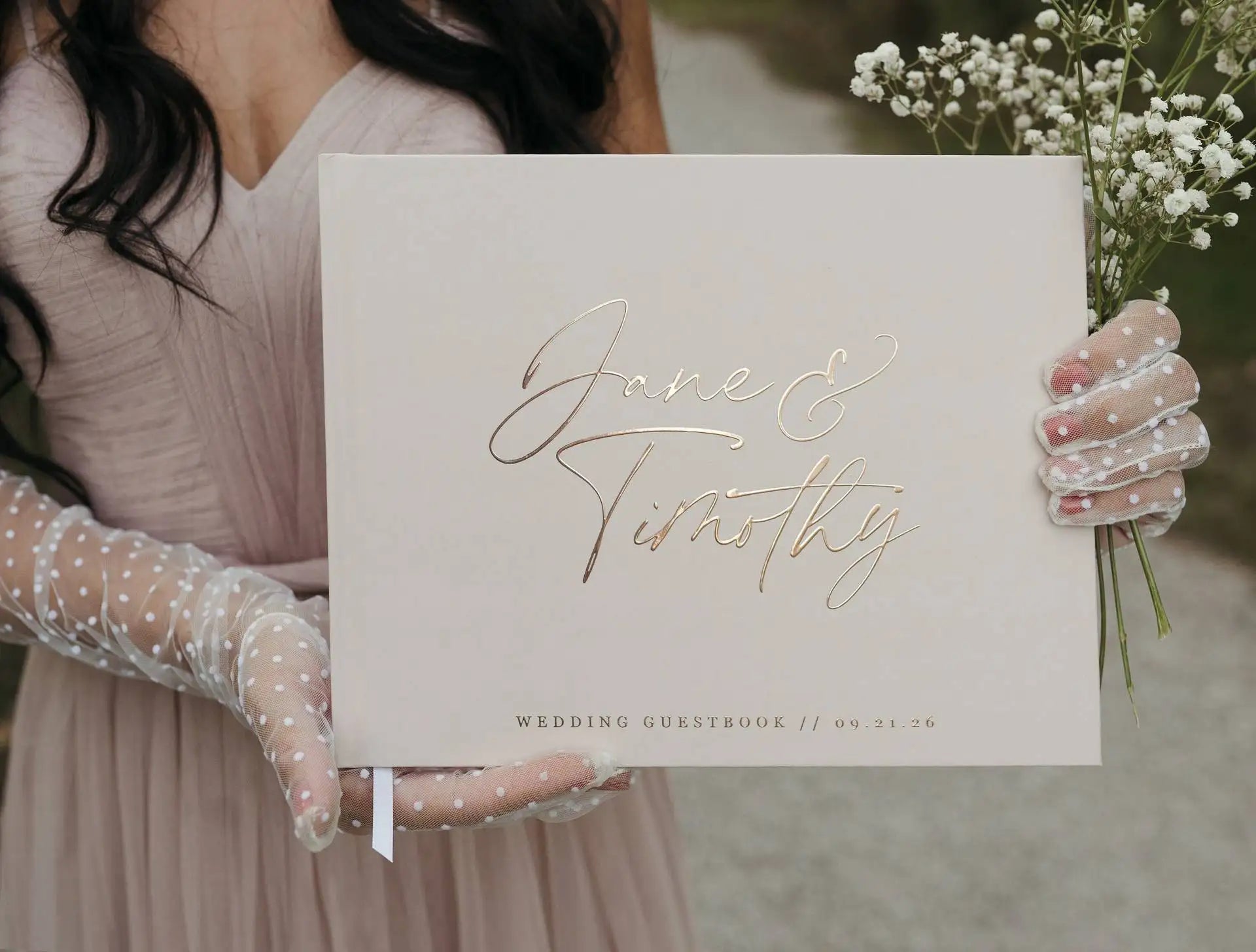 Woman in a blush dress and polka-dot gloves holds a wedding guestbook with "Jane & Timothy" in gold script, dated 09.21.26, alongside baby's breath flowers.
