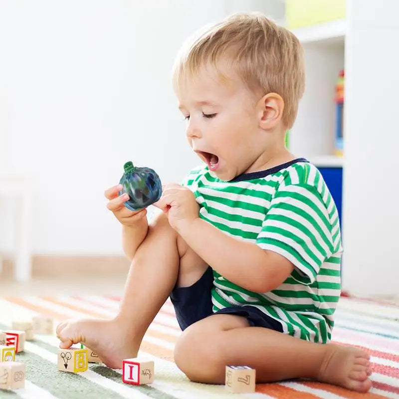 A toddler in a green striped shirt sits on a colorful rug, surprised while holding a small toy. Wooden blocks are scattered around on the floor.