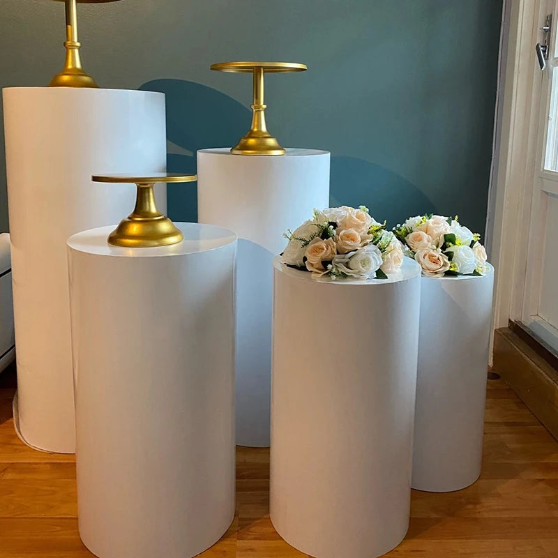 White cylindrical pedestals with gold cake stands and floral arrangements of cream roses and greenery, set against a blue wall in a bright room with wooden flooring.