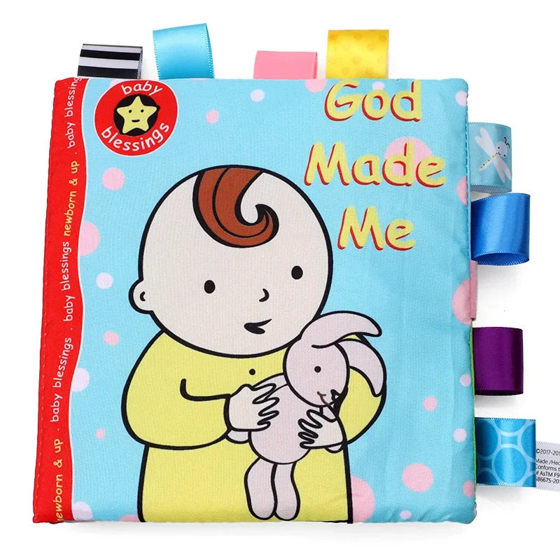Cloth baby book cover with a cartoon baby holding a rabbit. Text reads "God Made Me." The cover is blue with pink polka dots and colorful ribbon tabs on the edges.