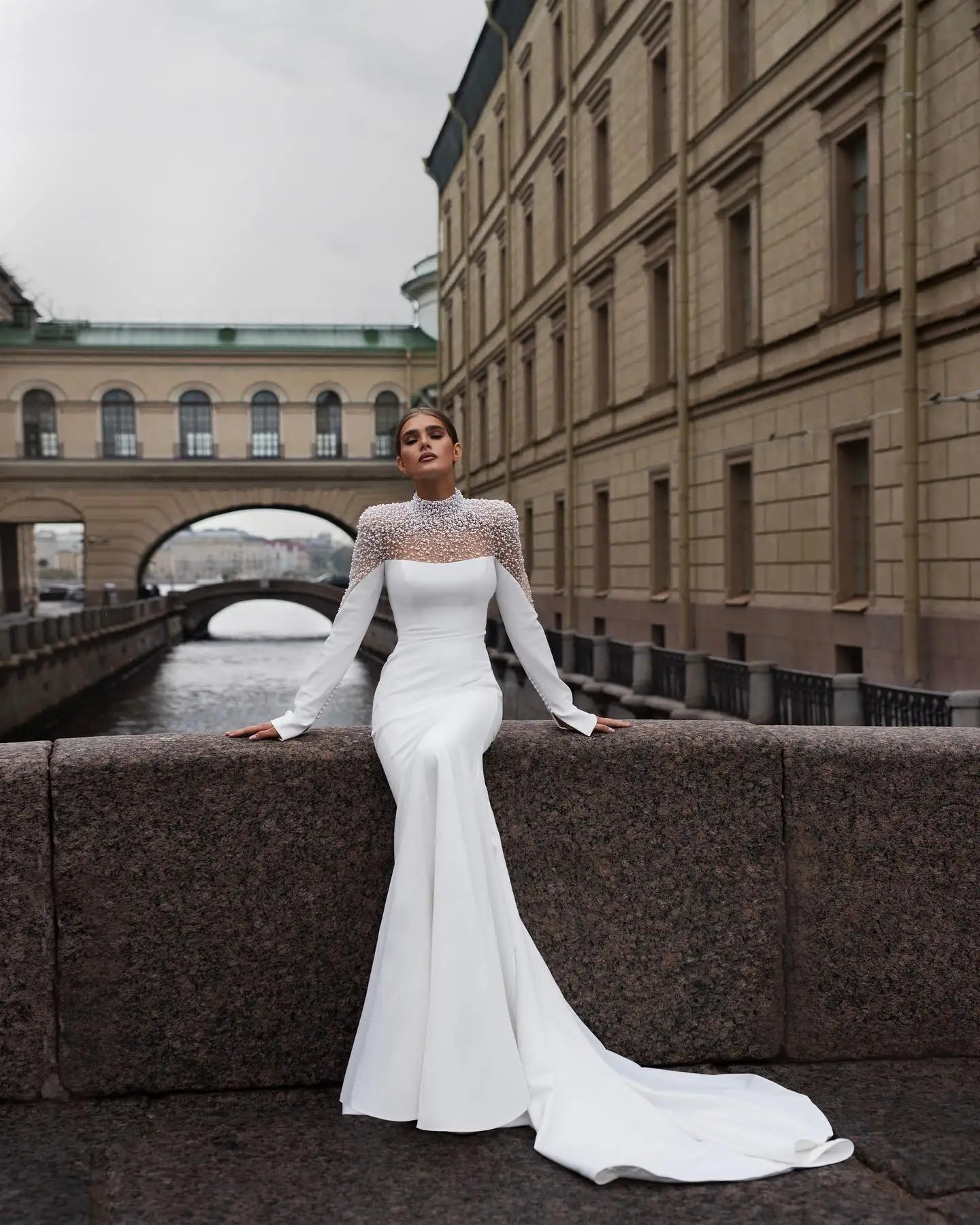 A woman in a sleek white gown with ornate sleeves leans against a stone wall by a canal. The backdrop features elegant, historic architecture and an arched bridge.