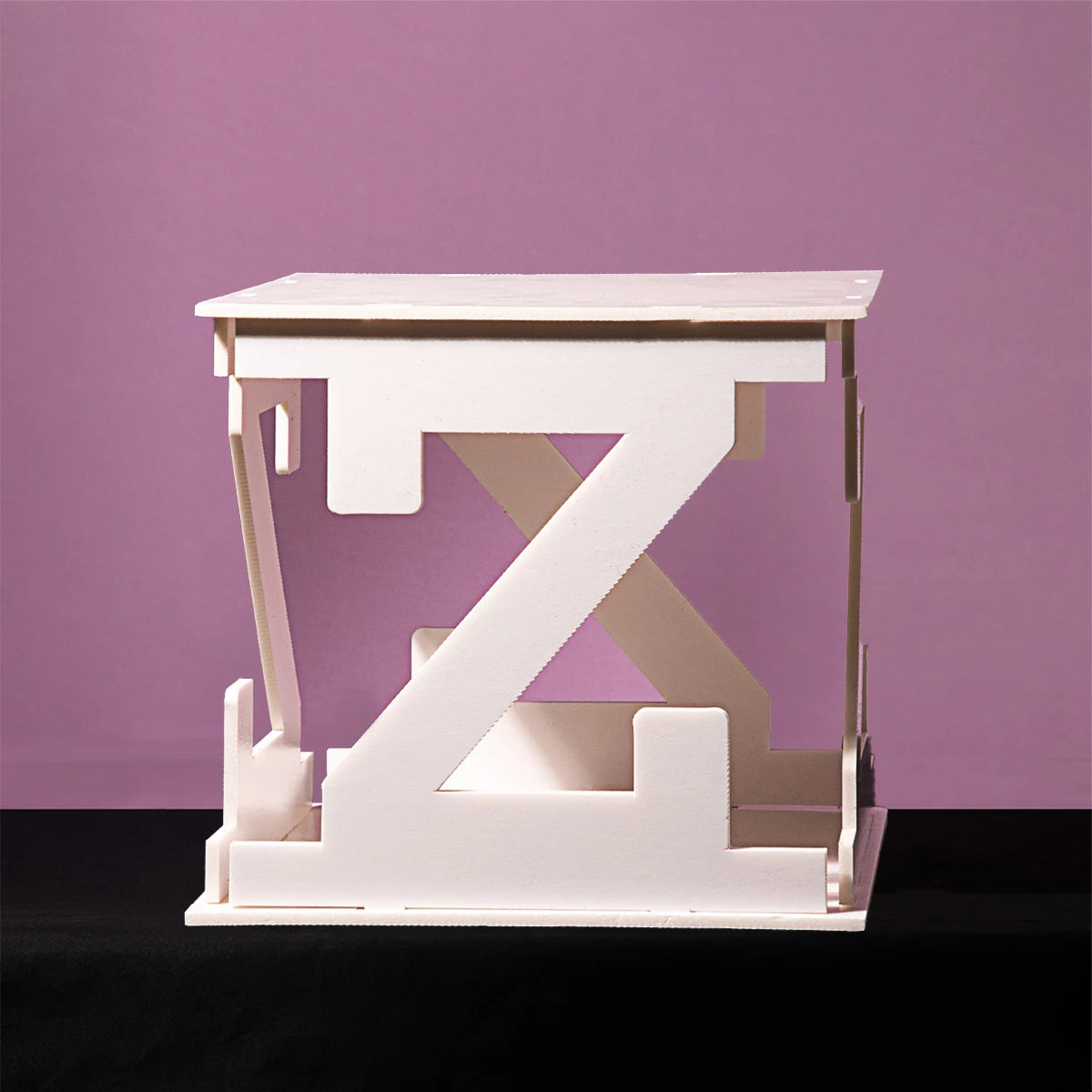 White geometric sculpture resembling a table with a large letter "Z" on the side, set against a purple background and placed on a black surface.