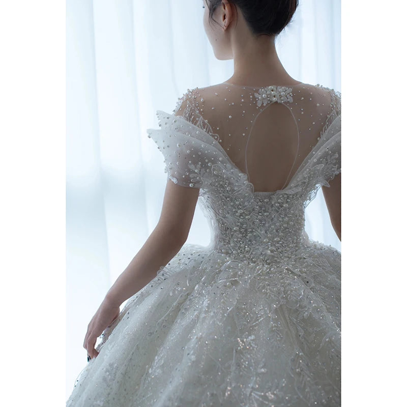 A woman in a white, intricately beaded wedding gown with a sheer back and cap sleeves stands softly lit by daylight against a plain background.