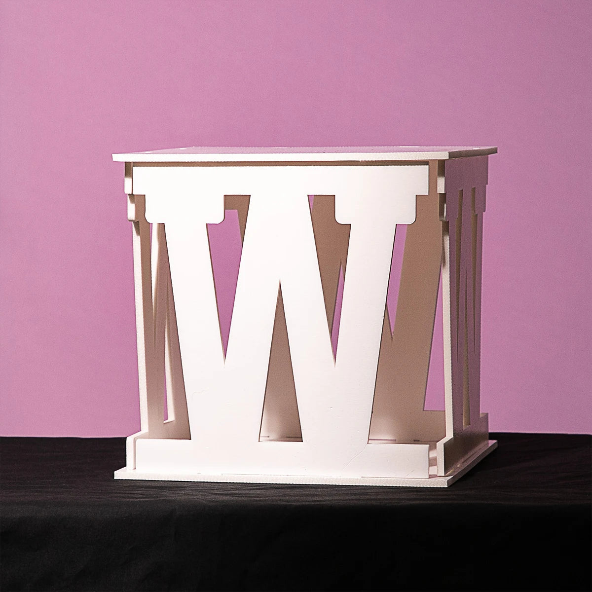 White cube-shaped table with cut-out "W" letters on all sides, set against a pink background and placed on a dark surface.