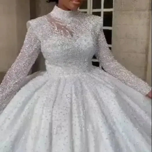 A woman wearing a sparkling, embellished white gown with long sleeves and a high neckline, featuring a full skirt and a sheer neckline detail.
