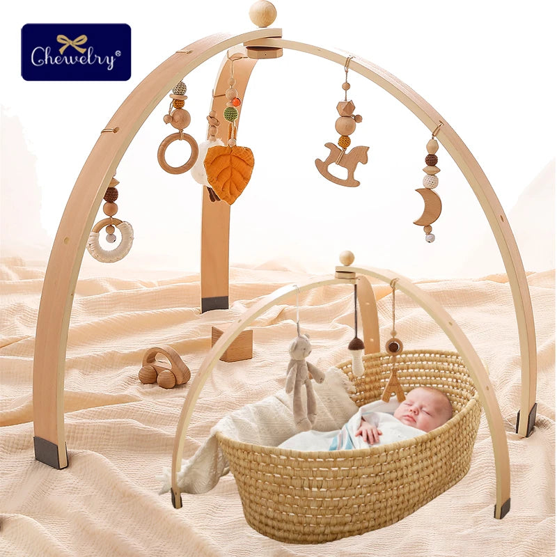 A baby sleeps in a wicker bassinet under a wooden play gym with hanging toys like a leaf, horse, and moon. The setup is on a soft, cream-colored blanket.